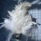 A white decorative feather ornament with layered soft feathers, a rhinestone-embellished black bead, iridescent ribbon, and white beads, photographed on a dark textured fabric background.