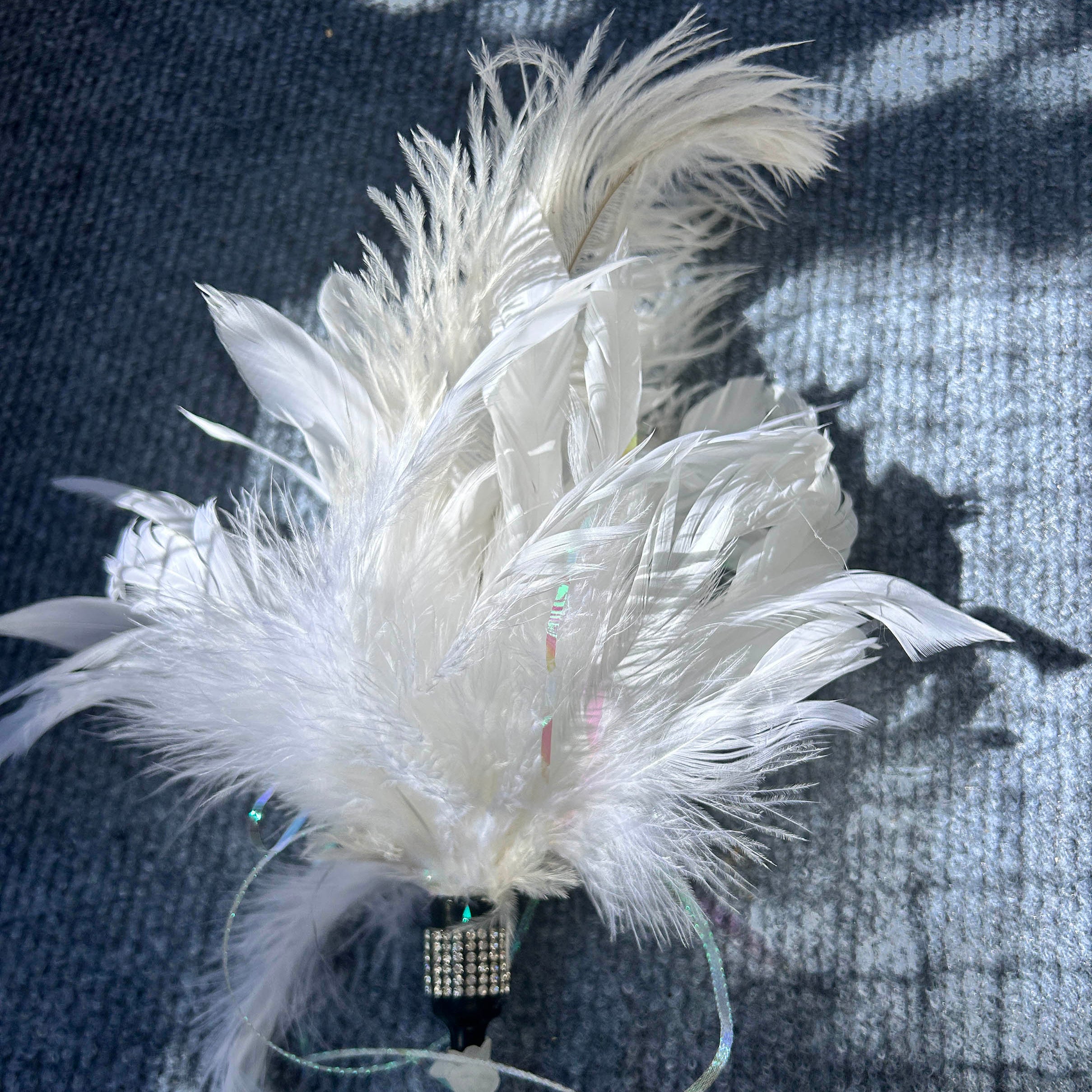 A white decorative feather ornament with layered soft feathers, a rhinestone-embellished black bead, iridescent ribbon, and white beads, photographed on a dark textured fabric background.