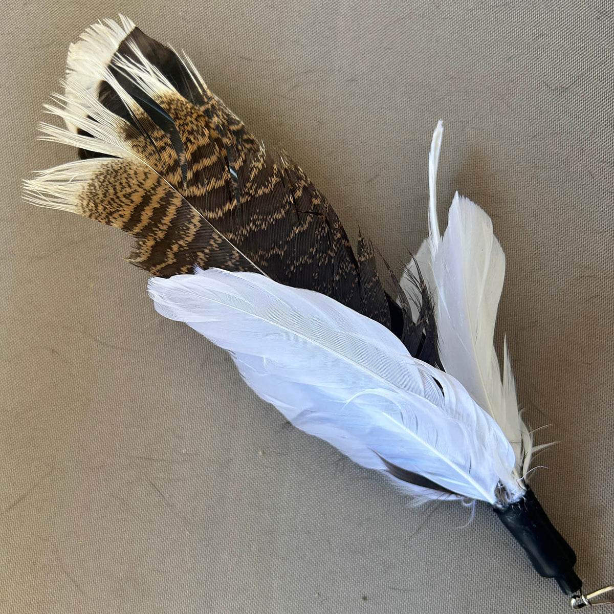 A decorative feather toy with a black wrapped handle and a cluster of feathers including white feathers and a brown-and-black patterned feather, placed on a beige textured surface.